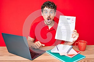 Young caucasian man with curly hair showing a passed exam sitting on the table scared and amazed with open mouth for surprise,