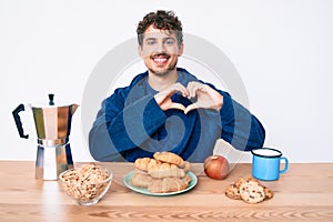 Young caucasian man with curly hair sitting on the table having breakfast smiling in love doing heart symbol shape with hands