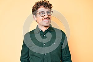 Young caucasian man with curly hair wearing casual clothes and glasses looking away to side with smile on face, natural expression