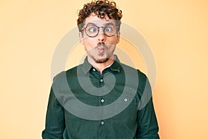 Young caucasian man with curly hair wearing casual clothes and glasses making fish face with lips, crazy and comical gesture