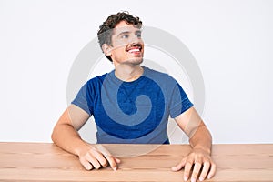 Young caucasian man with curly hair wearing casual clothes sitting on the table looking away to side with smile on face, natural