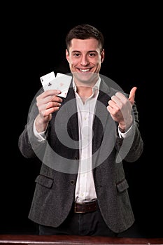 Young caucasian man wearing suit holding two aces in his hand and showing thumb up hand gesture with smile on black