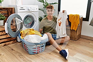 Young caucasian man doing laundry holding t shirt with stain looking positive and happy standing and smiling with a confident