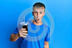 Young caucasian man drinking glass of soda beverage scared and amazed with open mouth for surprise, disbelief face