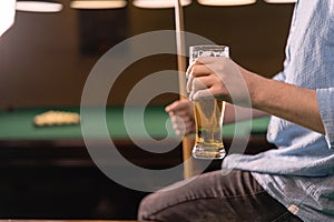 Young Caucasian man drinks beer while sitting on a billiard table, player resting after a game of billiards