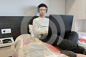 Young caucasian man enjoying music, laying on his bed with his laptop, smiling to the camera