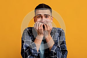 Young Caucasian man feeling scared, biting his nails in panic on orange studio background. Negative human emotions