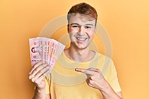 Young caucasian man holding 100 thai baht banknotes smiling happy pointing with hand and finger