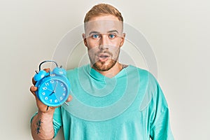 Young caucasian man holding alarm clock scared and amazed with open mouth for surprise, disbelief face