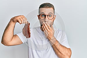 Young caucasian man holding beef steak covering mouth with hand, shocked and afraid for mistake