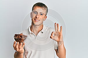 Young caucasian man holding bowl of star anise doing ok sign with fingers, smiling friendly gesturing excellent symbol