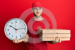 Young caucasian man holding delivery package and clock smiling looking to the side and staring away thinking