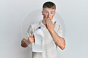 Young caucasian man holding detergent bottle covering mouth with hand, shocked and afraid for mistake