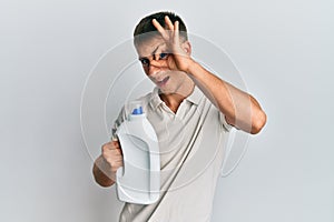 Young caucasian man holding detergent bottle smiling happy doing ok sign with hand on eye looking through fingers