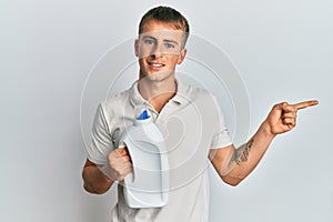 Young caucasian man holding detergent bottle smiling happy pointing with hand and finger to the side