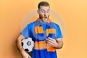 Young caucasian man holding football ball looking at smartphone afraid and shocked with surprise and amazed expression, fear and