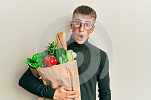 Young caucasian man holding paper bag with groceries scared and amazed with open mouth for surprise, disbelief face