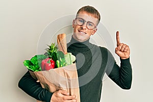 Young caucasian man holding paper bag with groceries surprised with an idea or question pointing finger with happy face, number