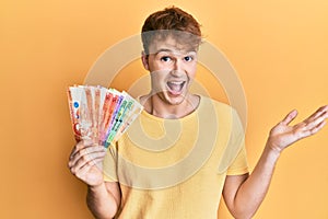 Young caucasian man holding philippine peso banknotes celebrating achievement with happy smile and winner expression with raised