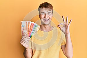 Young caucasian man holding philippine peso banknotes doing ok sign with fingers, smiling friendly gesturing excellent symbol