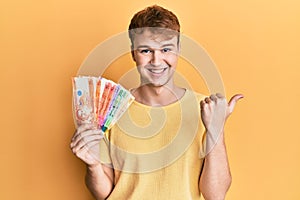 Young caucasian man holding philippine peso banknotes pointing thumb up to the side smiling happy with open mouth