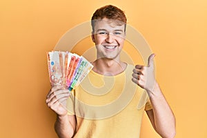 Young caucasian man holding philippine peso banknotes smiling happy and positive, thumb up doing excellent and approval sign