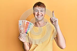 Young caucasian man holding philippine peso banknotes smiling with an idea or question pointing finger with happy face, number one