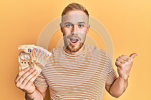Young caucasian man holding south african 20 rand banknotes pointing thumb up to the side smiling happy with open mouth