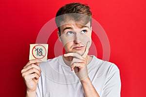 Young caucasian man holding under 18 prohibition sticker serious face thinking about question with hand on chin, thoughtful about