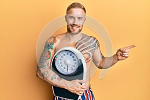 Young caucasian man holding weight machine to balance weight loss smiling happy pointing with hand and finger to the side