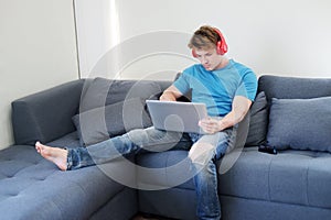 Young Caucasian man listening to musice by headphone and searching online social media with laptop in living room.