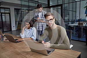 Young caucasian man, male office worker using laptop, sitting at office desk and looking at camera. Group of young