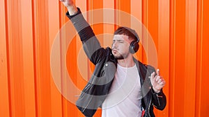 Young caucasian man in music headphones listens to music and dances on orange background, prance, slow motion