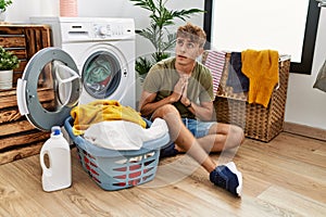 Young caucasian man putting dirty laundry into washing machine begging and praying with hands together with hope expression on