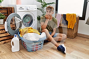 Young caucasian man putting dirty laundry into washing machine looking stressed and nervous with hands on mouth biting nails