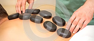 Young caucasian man receiving back massage with black stones by masseur in spa salon. A woman getting a spa treatment.