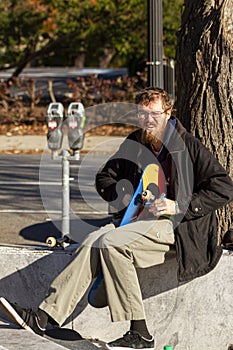 young caucasian man with red goatee beard is sitting on a wall assembling his skateboard.