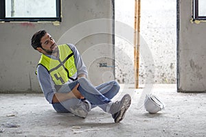 Young caucasian man in safety vest gripped his knee and screams in pain. Due to injuries at the construction site