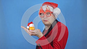 A young caucasian man in a santa claus costume holds a mandarin in his hand and looks at the camera. Blue background