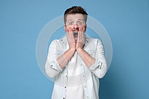 Young caucasian man in shock opening mouth and touching face with hands, looking in full disbelief