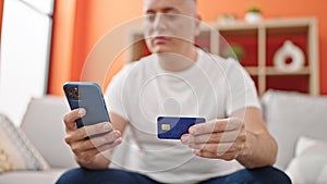 Young caucasian man shopping with smartphone and credit card sitting on sofa at home