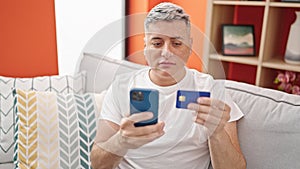 Young caucasian man shopping with smartphone and credit card sitting on sofa at home