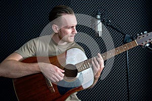 Young caucasian man sings while playing an acoustic guitar in front of black soundproofing walls. Musicians producing music in