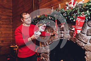 Young caucasian man sitting at fireplace wearing sweater holding mug near decorated christmas tree.