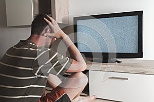 Young caucasian man sitting with head in hands looking to grey graining television. Depression, psychic, mentally ill concept