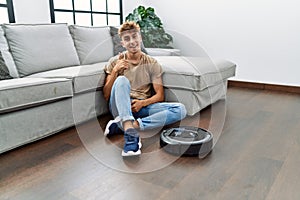 Young caucasian man sitting at home by vacuum robot smiling happy and positive, thumb up doing excellent and approval sign