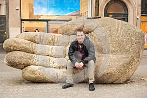 Young caucasian man sitting on huge stone hand on street of Antwerp, Belgium in early spring day. Antwerpen travel tourism