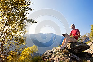 Young caucasian man sitting outdoor on a rock working on a laptop pc in mountain area.