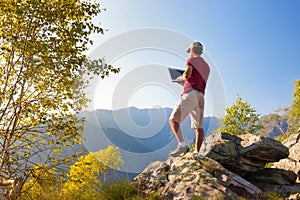 Young caucasian man sitting outdoor on a rock working on a laptop pc in mountain area.