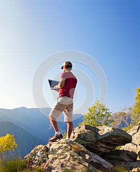 Young caucasian man sitting outdoor on a rock working on a laptop pc in mountain area.